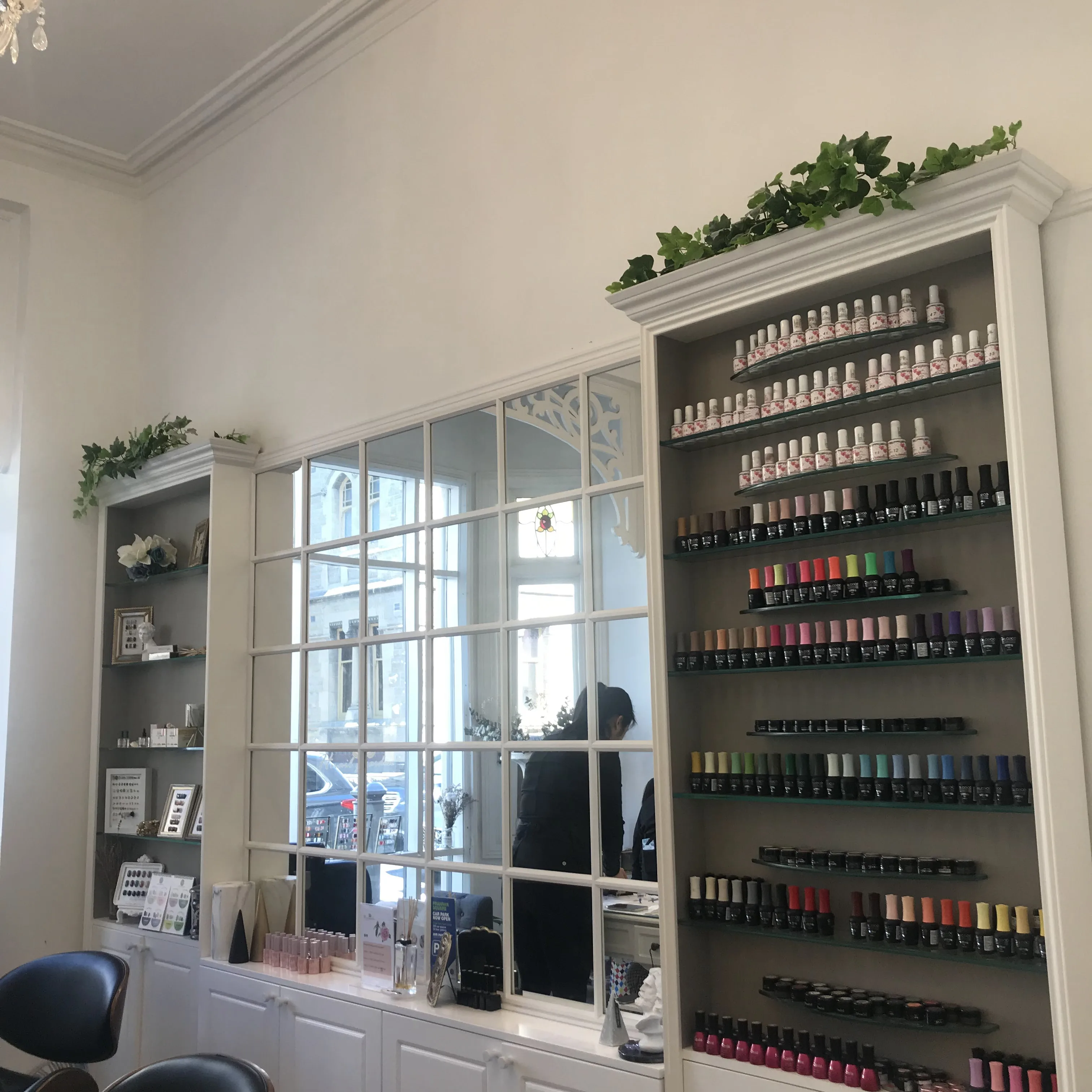 The Prahran salon interior — a floor-to-ceiling wall of gel-polish bottles arranged by colour, beneath an arched grille window with trailing ivy